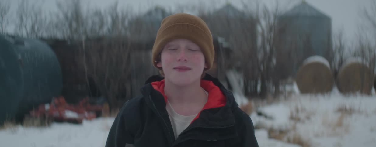A portrait of a young boy with freckles and reddish hair in a beanie, standing outside in the snow on a farm