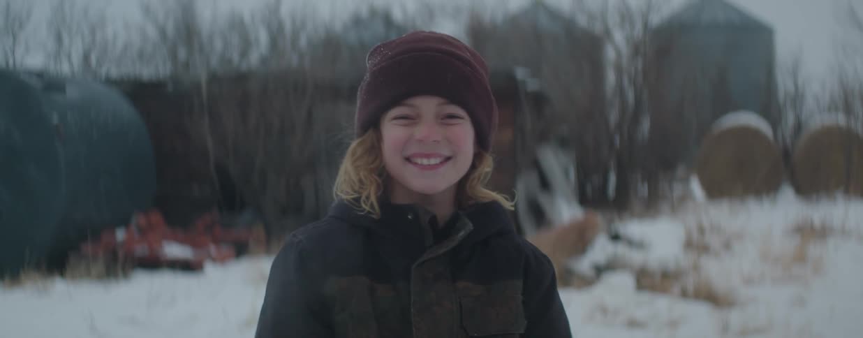 A happy young boy wearing a winter beanie and jacket smiles genuinely at the camera in a snowy rural landscape