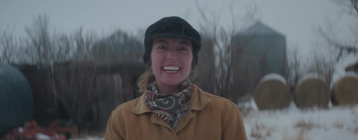 A joyful female farmer warmly dressed in a winter hat and coat smiles and laughs in the falling snow on her farm