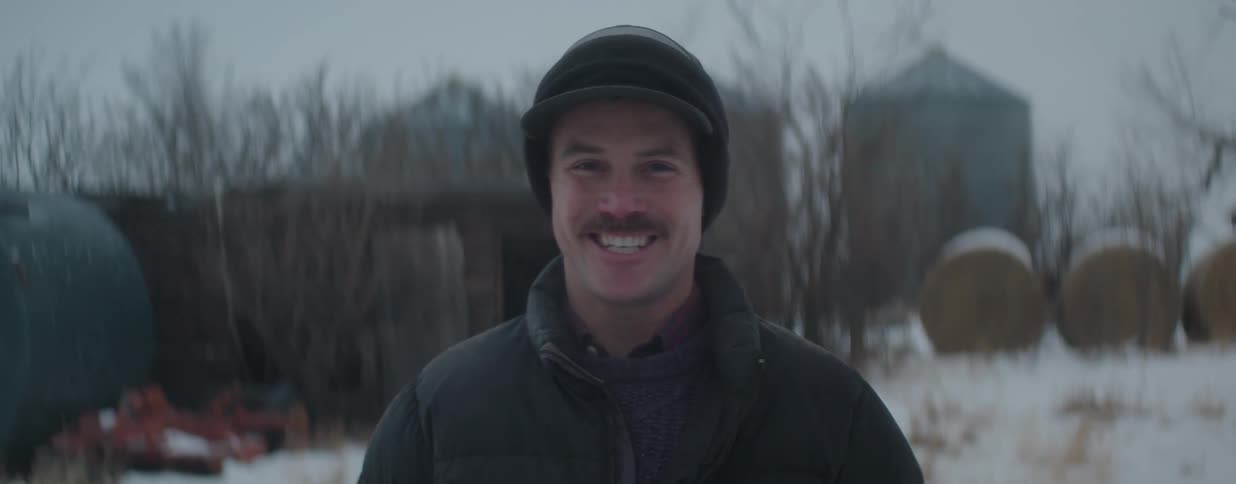 Close-up portrait of a mustached man in winter clothing smiling warmly in a snowy, rural farm setting