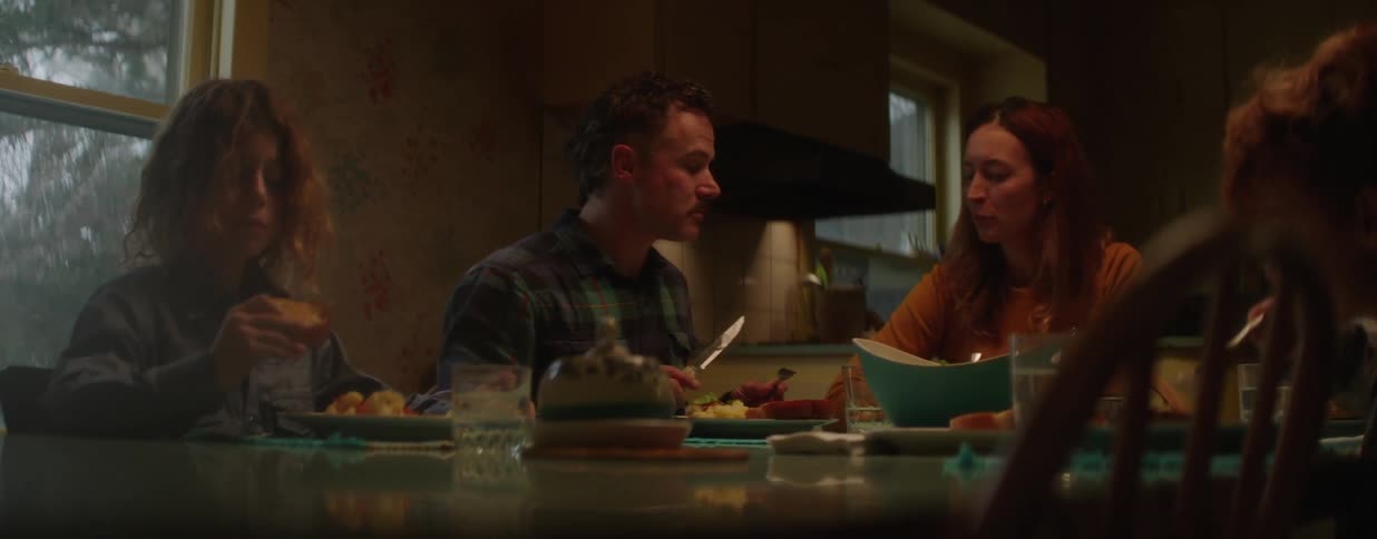 A family of three eats dinner and has a conversation while sitting at a dining table in their dimly lit kitchen at night