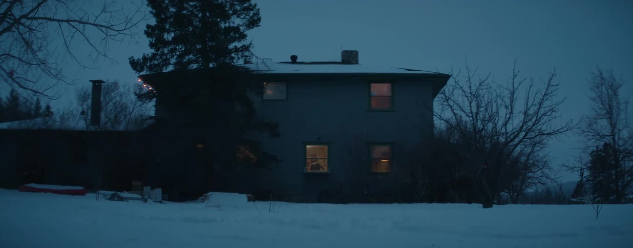 Warm lights glow from the windows of a quiet house on a cold, snowy winter evening