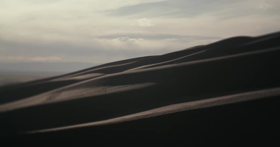 The rolling silhouettes of dark sand dunes are cast in shadow under a soft, hazy sky