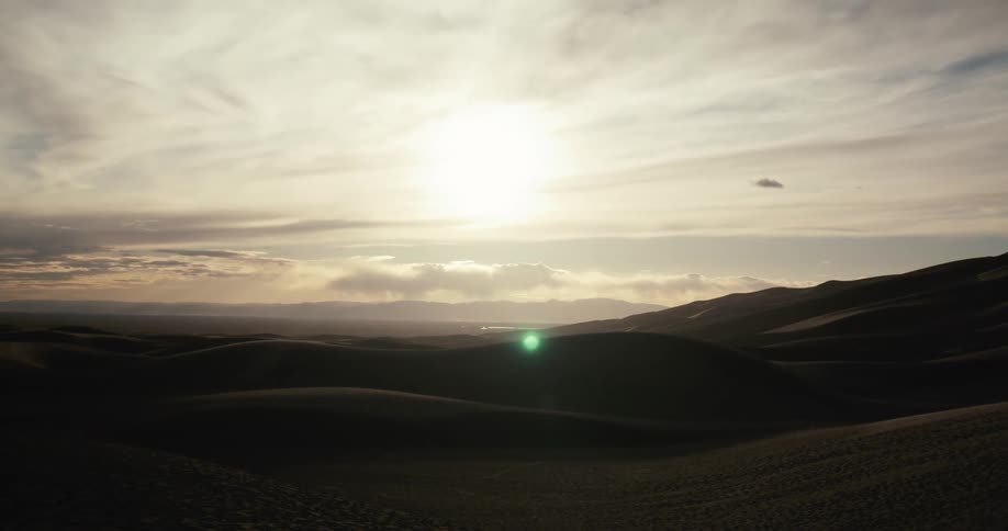 A reveal shot from behind a person's silhouette shows a vast landscape of rolling desert sand dunes at sunset