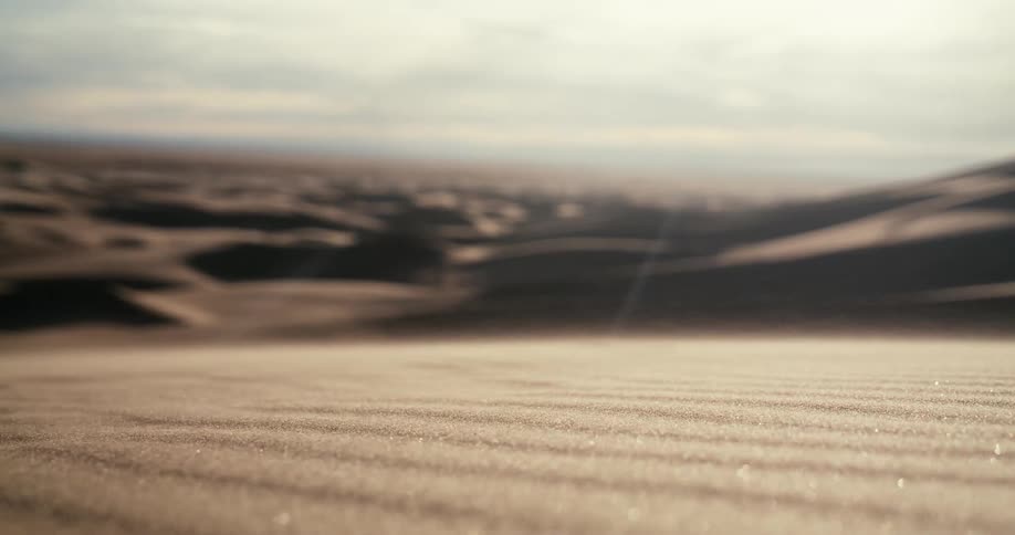 A shallow focus close-up captures wind blowing sparkling sand grains across the surface of a vast desert dune