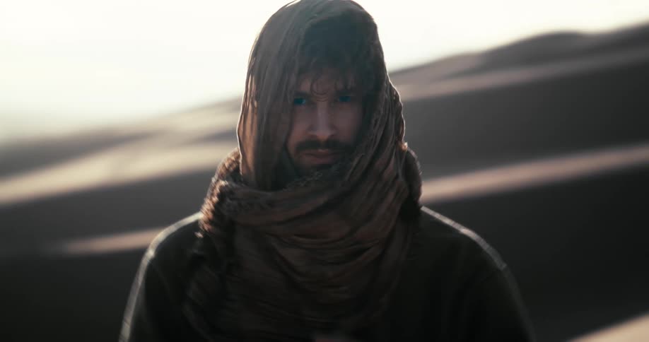 A mysterious man with piercing blue eyes stares intently before covering his face with a scarf in the desert