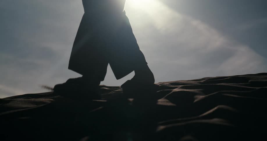 A dramatic slow motion low-angle shot of a person's silhouette walking through the sand against a bright sun