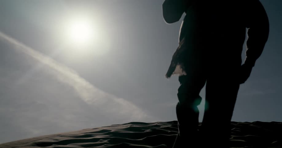 A low-angle slow-motion silhouette of a person walking up a sand dune with their scarf blowing in the wind