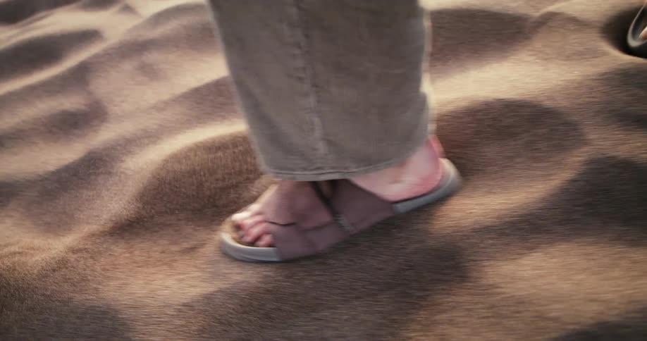 A slow-motion close-up of a person's feet in sandals walking through rippling desert sand
