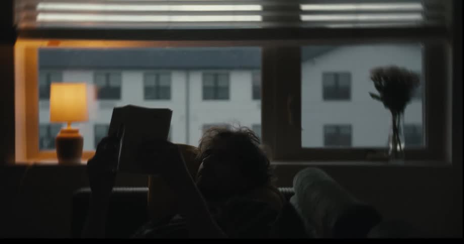 A silhouetted person reads a book while relaxing on a couch by a large window on a gloomy day