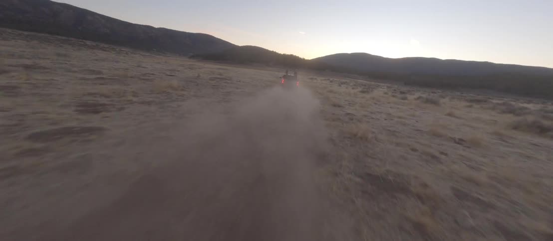 An aerial shot follows a pickup truck with two people in the back as it kicks up dust while off-roading through a field at sunset