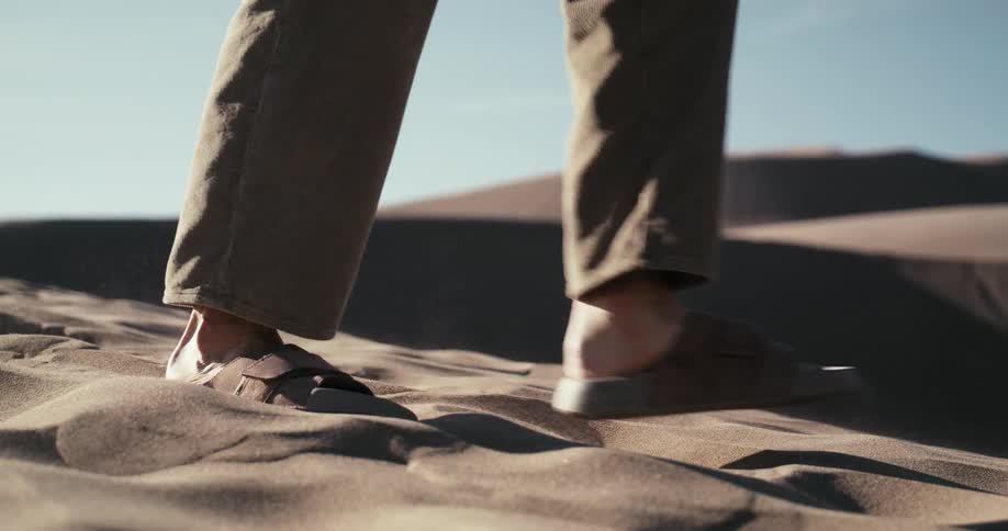 A person's feet in sandals walk through the soft desert sand in a dramatic slow-motion close-up