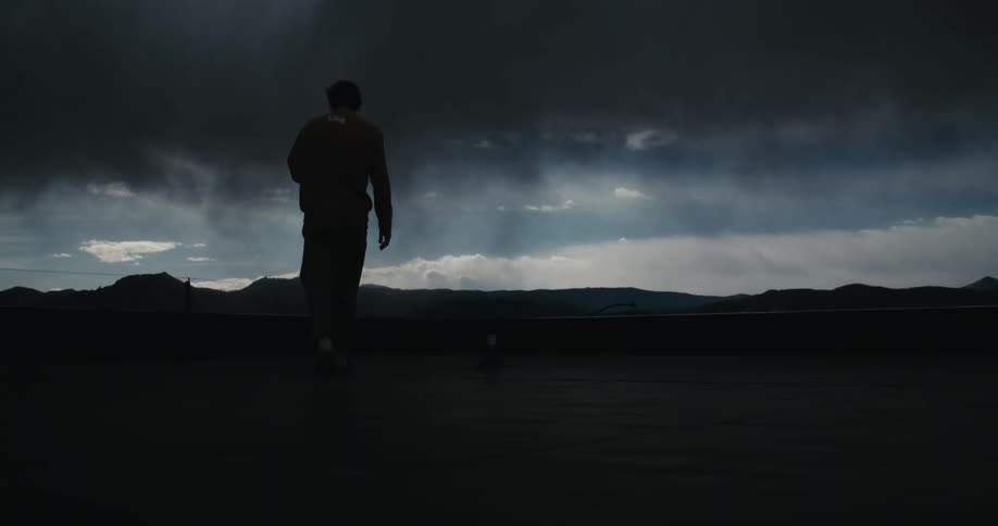 In slow motion, a silhouetted man walks towards a dramatic, stormy sky over a mountain range