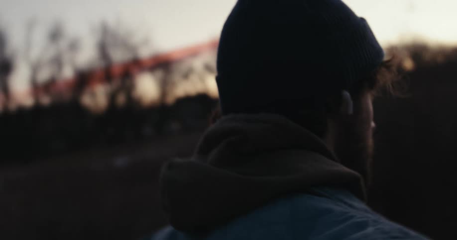 A thoughtful young man wearing a beanie and wireless earbuds looks into the distance while walking outdoors at sunset