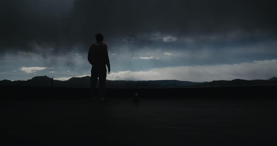 A silhouetted man stands alone on a rooftop overlooking a dramatic, stormy sky and distant mountains