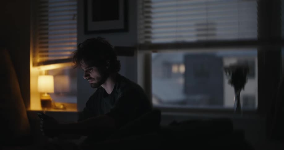 A stressed man scrolls on his phone late at night, the screen's glow illuminating his face in the dark room