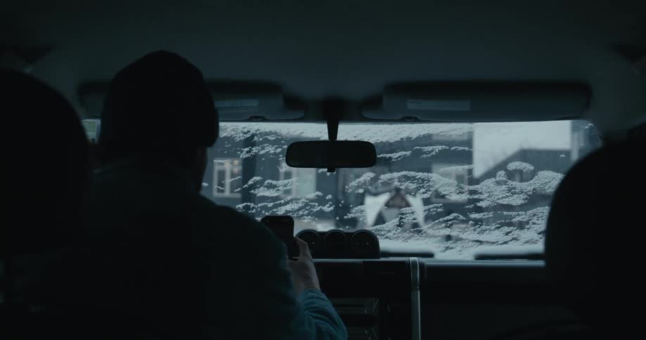 A silhouetted man in a beanie sits in a parked car and uses his smartphone, with a view of a snowy street through the front windshield