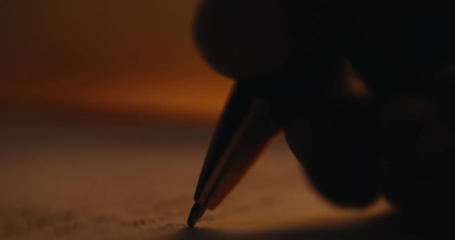 A close-up macro shot of a silhouetted hand writing a letter with a pen on paper by warm candlelight