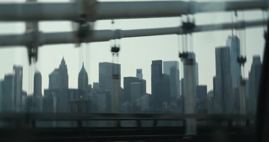 A cinematic tracking shot from a moving vehicle reveals the iconic New York City skyline while crossing a bridge on an overcast day
