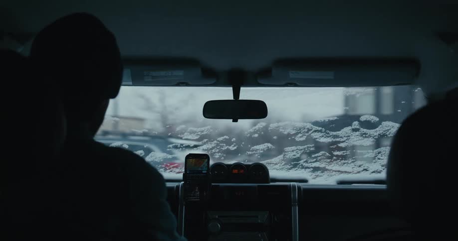 A moody, over-the-shoulder shot from inside a vehicle driving through a snowy landscape on a cold winter day