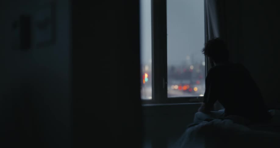 A pensive person sits in silhouette on a bed, gazing out a window at the bustling city lights below