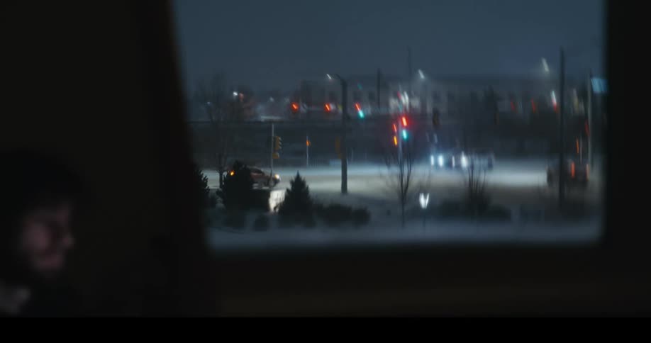 A lonely young man sits in a dark room, staring out the window at the blurry traffic lights on a cold, snowy night