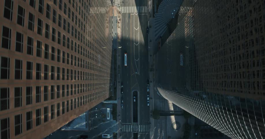A dramatic top-down aerial view slowly ascends over a sunlit street canyon carved by towering skyscrapers in a modern city