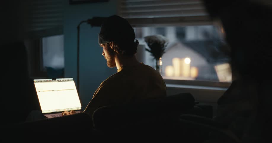 A young man wearing a beanie works on his laptop in a dimly lit room at night