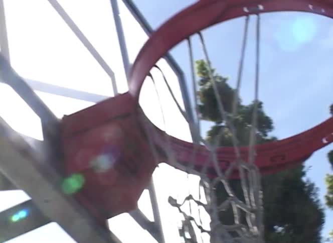 A basketball flies through the air and scores a basket on a sunny outdoor court, filmed from a low angle with lens flare