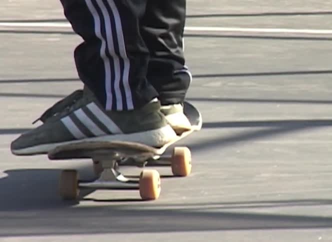 A slow zoom focuses on the shoes and skateboard of a person standing on an outdoor basketball court