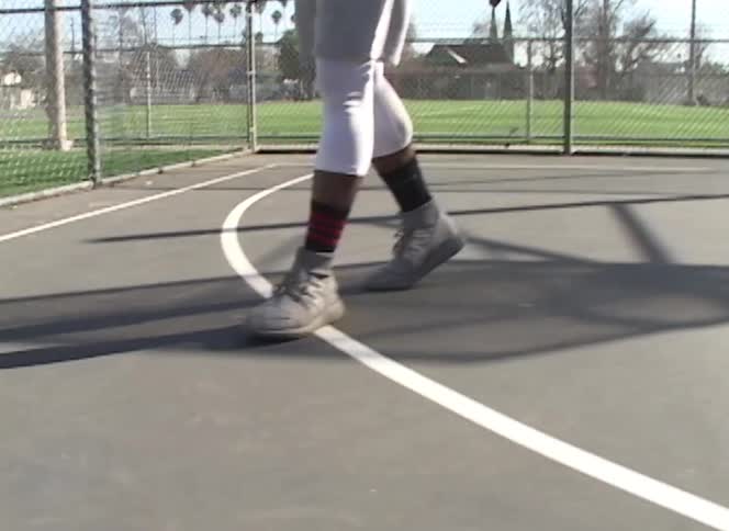 Low-angle tracking shot of a man's athletic shoes as he walks across a sunny outdoor basketball court