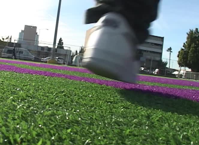 Low angle shot of a person in white sneakers walking across a green and purple artificial turf field on a sunny day