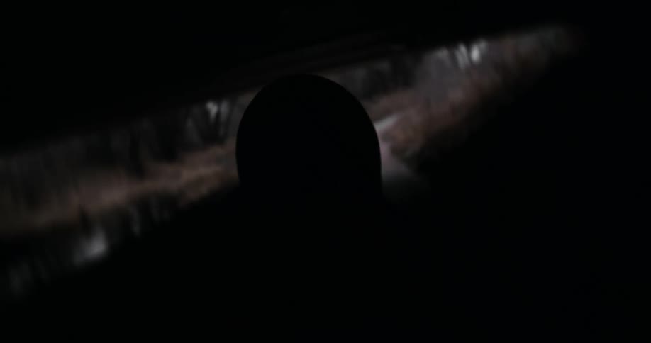 An over-the-shoulder slow-motion shot follows a rider with a helmet down a paved trail through a bleak landscape, passing under a bridge