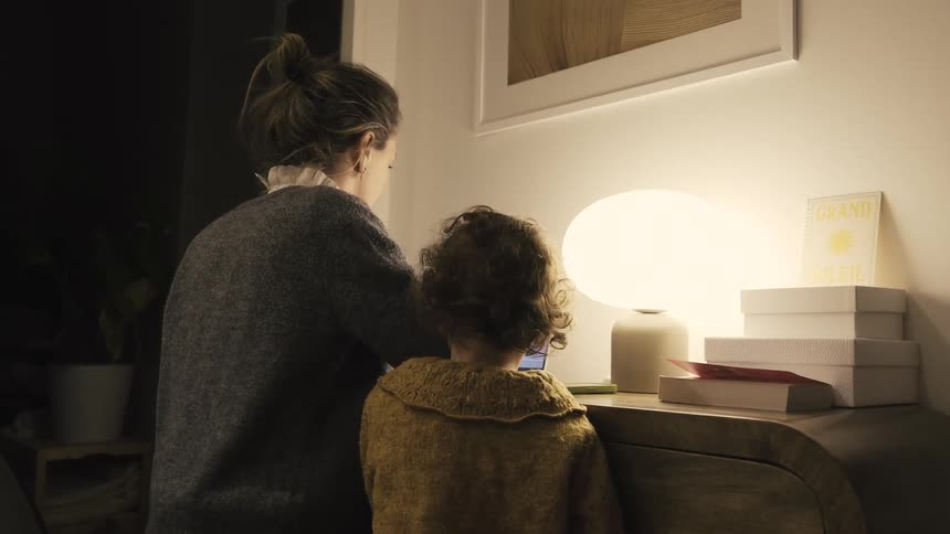 A mother and her young child share a quiet moment watching a laptop together by the warm glow of a lamp at home