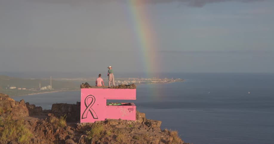A couple admires a vibrant rainbow over the ocean from a pink bunker painted with a breast cancer awareness ribbon on a cliffside