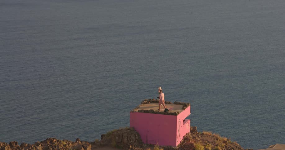A man and woman take photos on a brightly painted pink bunker perched on a cliff overlooking the ocean