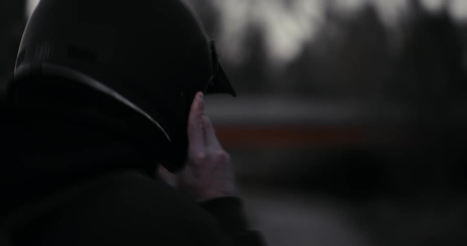 A mysterious man seen from behind puts on a black helmet and pulls up his hood in a moody, out-of-focus outdoor setting
