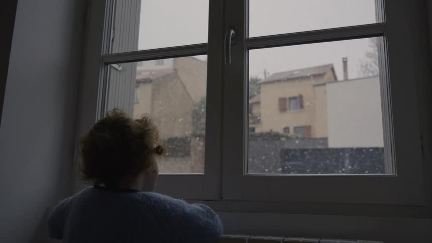 A small child with curly hair stands by a window, watching snow gently fall outside