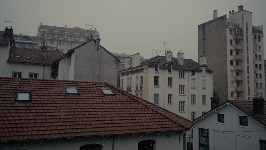 Slow-motion snow falls over the rooftops of a European city on a cold, overcast day