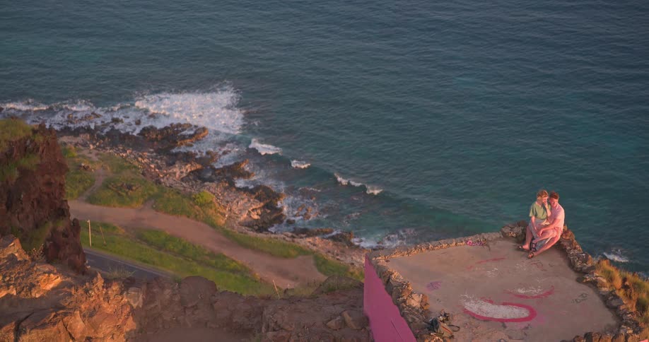 From a high angle, a father and his son sit on a pink concrete overlook, enjoying the expansive ocean view as the camera tilts down to reveal the coastline