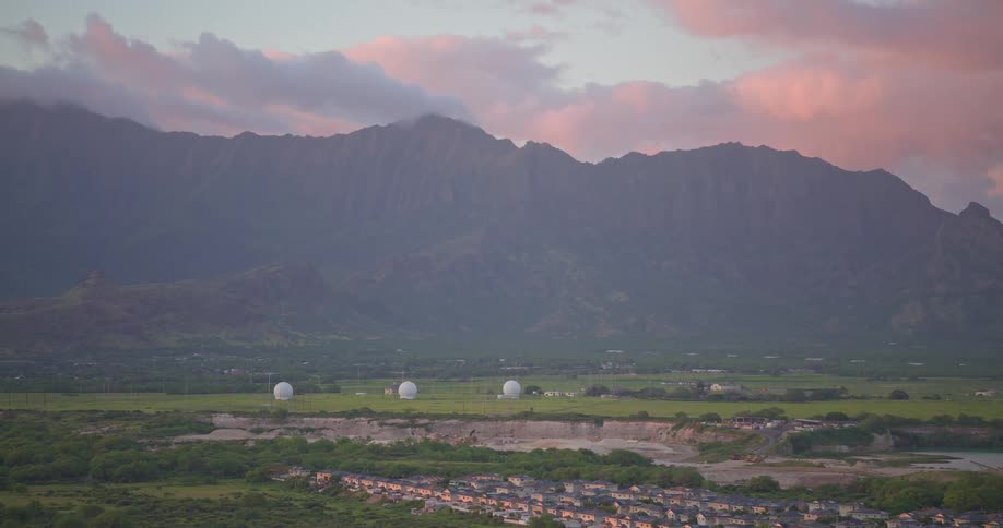 An aerial view pulls back to reveal a suburban development in a vast green valley with radar domes at the base of a Hawaiian mountain range at sunset
