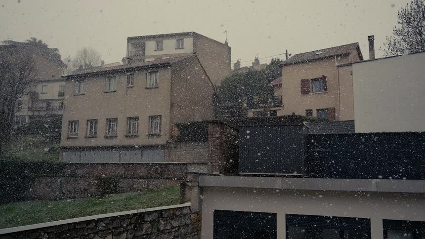 Heavy snow falls gently over the rooftops of a quiet residential neighborhood on an overcast day