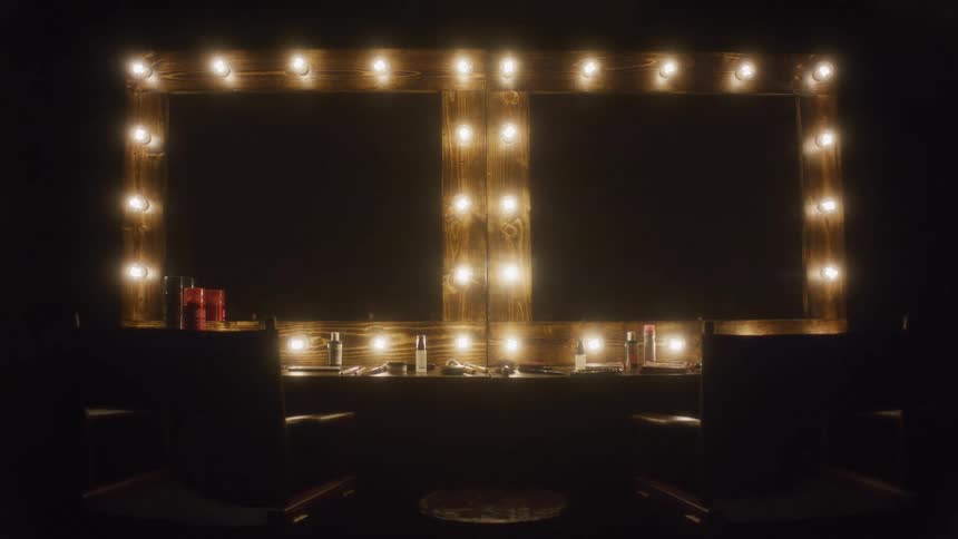 Slowly pushing into an empty, atmospheric backstage makeup station with two mirrors framed by warm, glowing bulbs