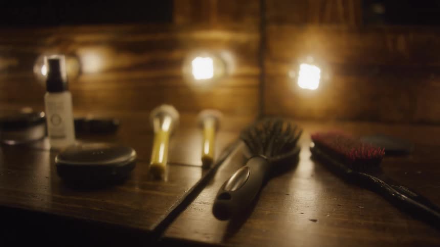 A slow rack focus reveals makeup brushes and cosmetics on a rustic wooden vanity table lit by warm bulbs