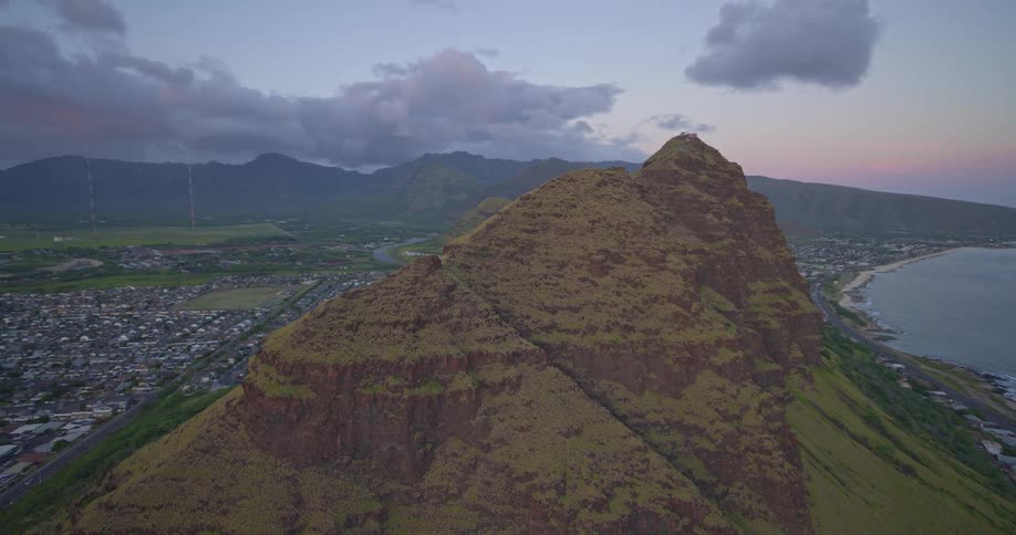 An aerial view slowly approaches a majestic coastal mountain in Hawaii at sunset, with a town and valley on one side and the ocean on the other