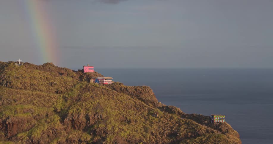 An aerial view slowly pulls back to reveal a vibrant rainbow over colorful, graffiti-covered bunkers on a rugged coastal cliff overlooking the ocean