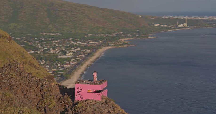Aerial shot orbits around a couple standing on a pink pillbox on a mountain ridge, revealing the vast ocean and coastal town below