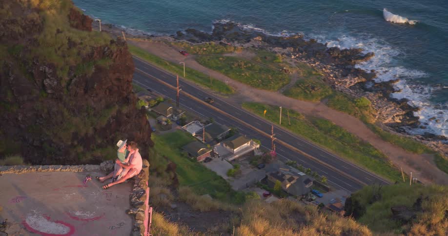 An aerial shot slowly pulls back from a romantic couple embracing on the edge of a cliff overlooking the ocean and a coastal highway at sunrise