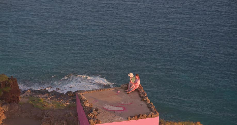 An aerial view reveals a couple sitting together on a cliffside lookout, sharing a quiet moment while overlooking the vast blue ocean