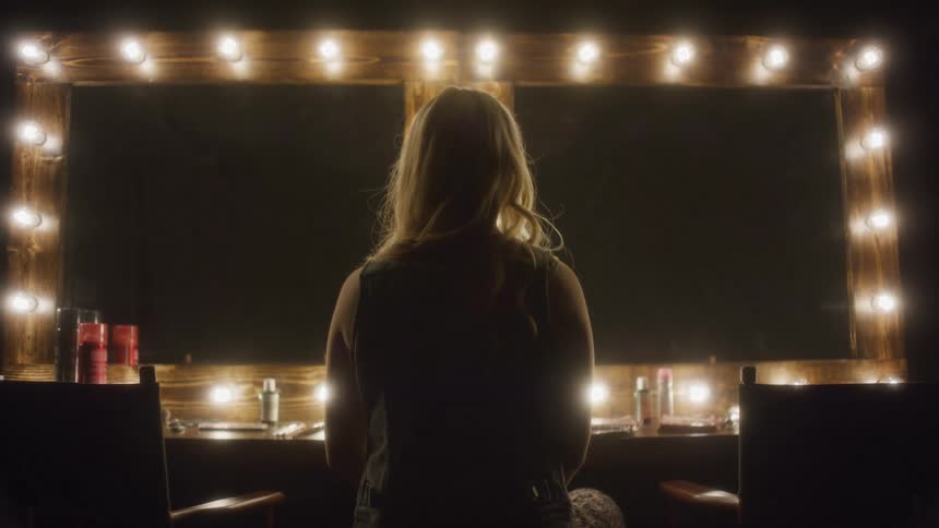 A woman sits in a dark dressing room, her back to the camera as it slowly pushes in on her and the lit vanity mirror